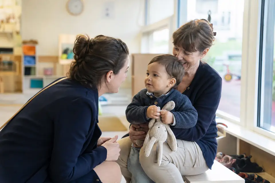 Une mère accroupie dans une salle de crèche dit au revoir à son fils d’un an, assis avec son doudou sur les genoux d’une auxiliaire souriante.