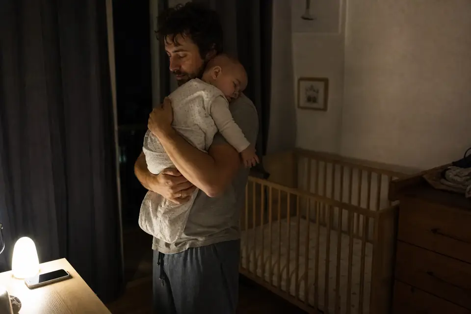 Un père berce son bébé de quatre mois endormi contre son épaule, dans une chambre à la lumière tamisée, à côté d’un lit à barreaux vide.