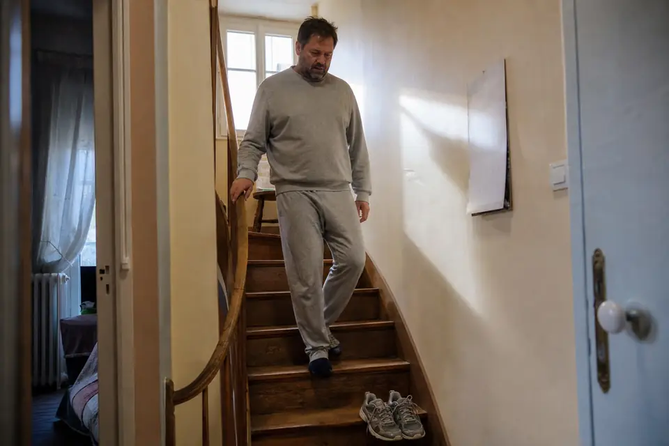 Un homme d’une cinquantaine d’années descend prudemment un escalier en bois en se tenant à la rampe, les jambes raides.