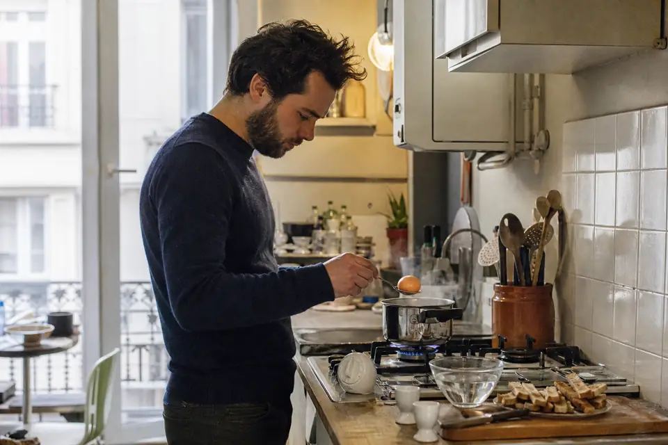 Un homme dépose un œuf avec une cuillère dans une petite casserole d’eau bouillante, à côté de coquetiers et de mouillettes.
