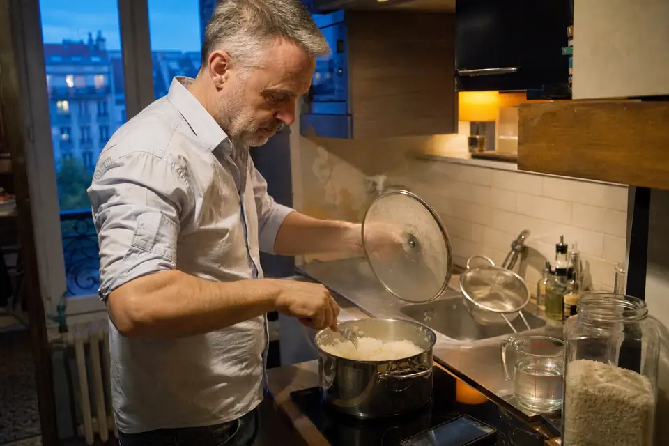 Un homme soulève le couvercle d’une casserole de riz basmati fumant et l’égrène à la fourchette, dans une petite cuisine d’appartement.