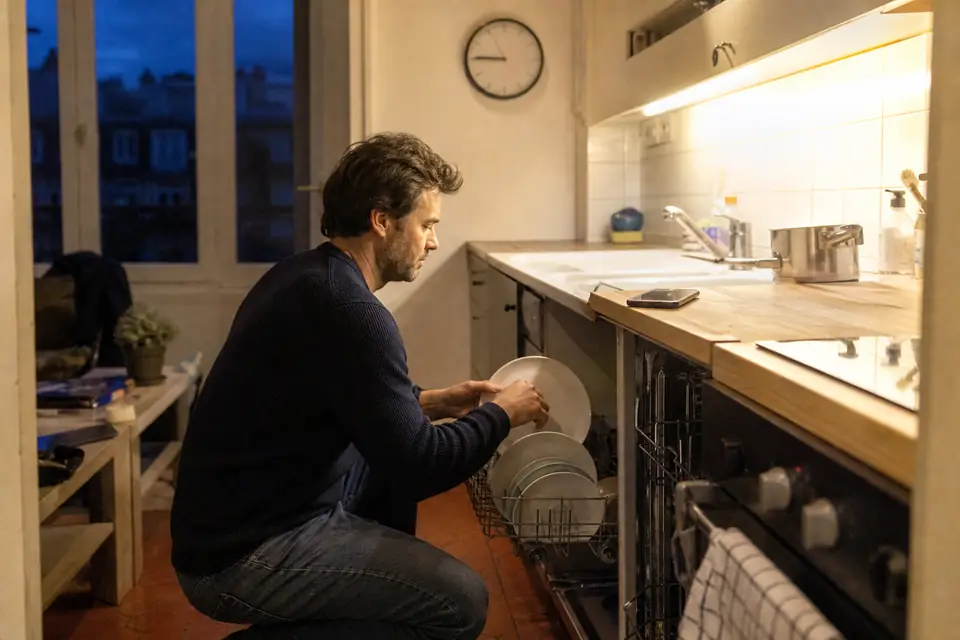 Un homme accroupi devant un lave-vaisselle ouvert glisse les dernières assiettes dans le panier du bas, un soir, dans une petite cuisine.