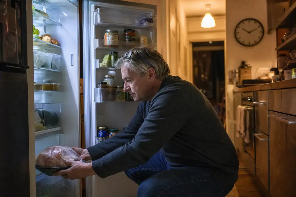 Un homme pose un poulet entier congelé, dans son emballage givré, sur une assiette creuse en bas d’un réfrigérateur ouvert.