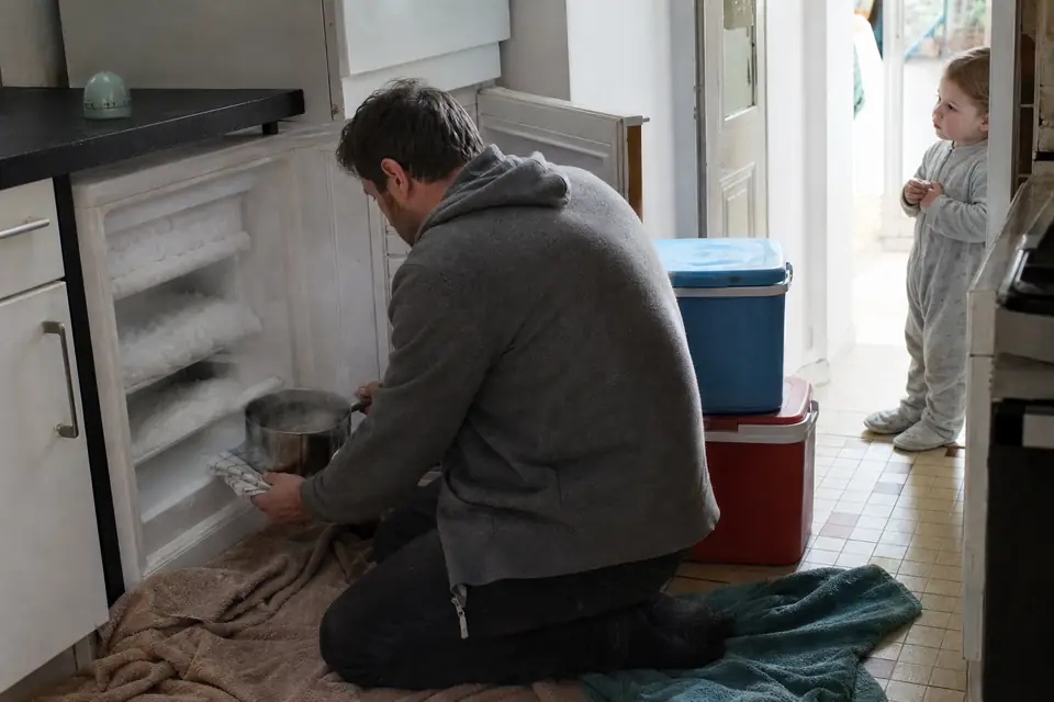 Un homme agenouillé devant un congélateur armoire ouvert et couvert de givre y dépose une casserole d’eau fumante, serviettes étalées au sol.