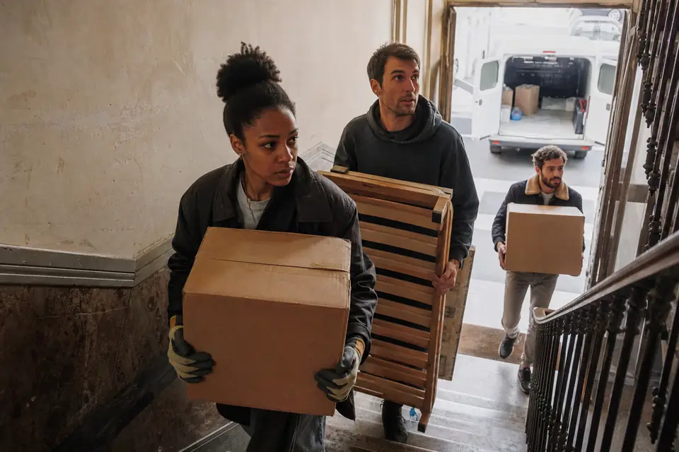 Trois amis descendent des cartons et un sommier démonté dans l’escalier d’un vieil immeuble, une camionnette de location garée devant la porte.