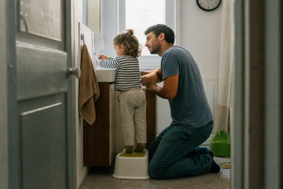 Un père agenouillé dans une petite salle de bains aide sa fille de deux ans et demi à se laver les mains, un pot vert posé dans un coin.