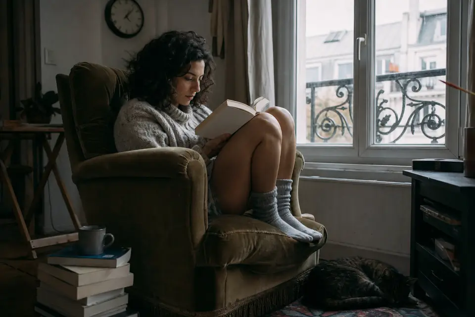 Une jeune femme lit un épais roman de poche, installée dans un fauteuil près d’une fenêtre, une tasse de thé posée sur une pile de livres.