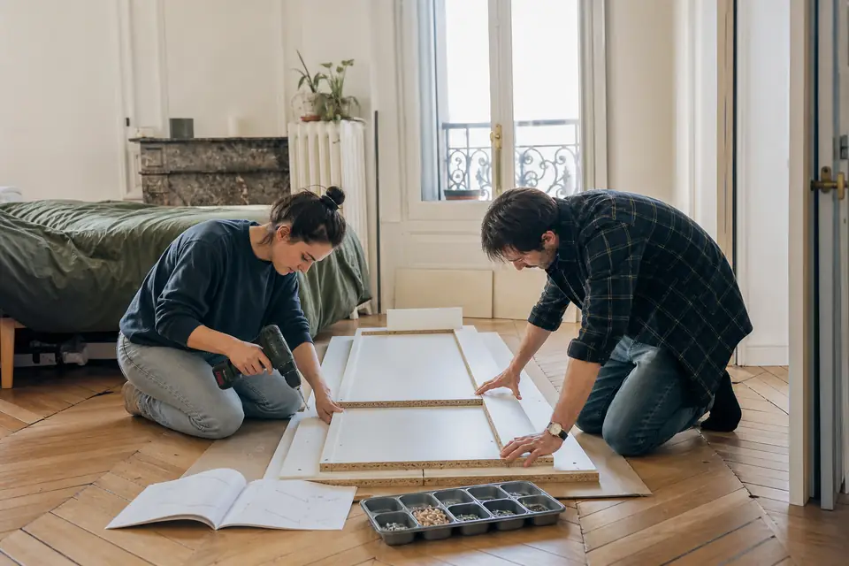 Deux personnes agenouillées sur des cartons dépliés assemblent le caisson d’une armoire blanche posé à plat sur le sol d’une chambre.