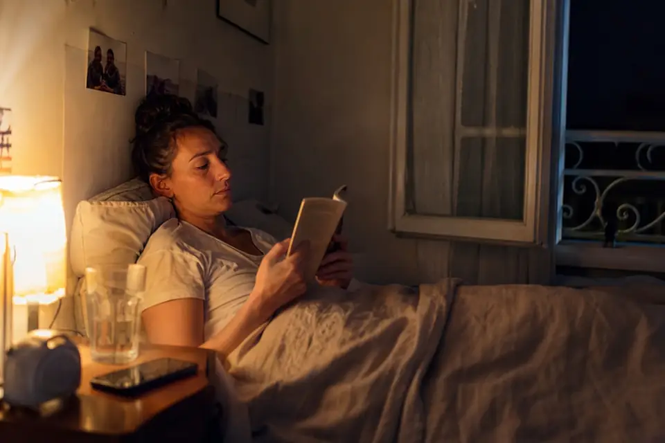 Une femme lit un livre au lit sous la lumière chaude d’une lampe de chevet, son téléphone posé écran contre la table de nuit.