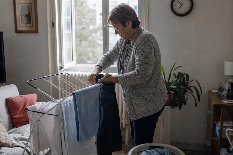 Une femme étend des serviettes et un jean sur un étendoir pliant dans un petit séjour, près d’une fenêtre entrouverte, un panier de linge à ses pieds.