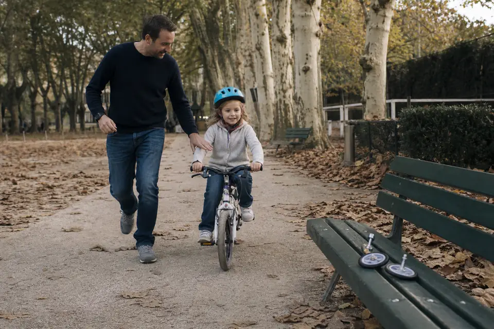 Un père court à côté de sa fille casquée qui pédale seule sur un petit vélo sans petites roues, dans une allée de parc en automne.
